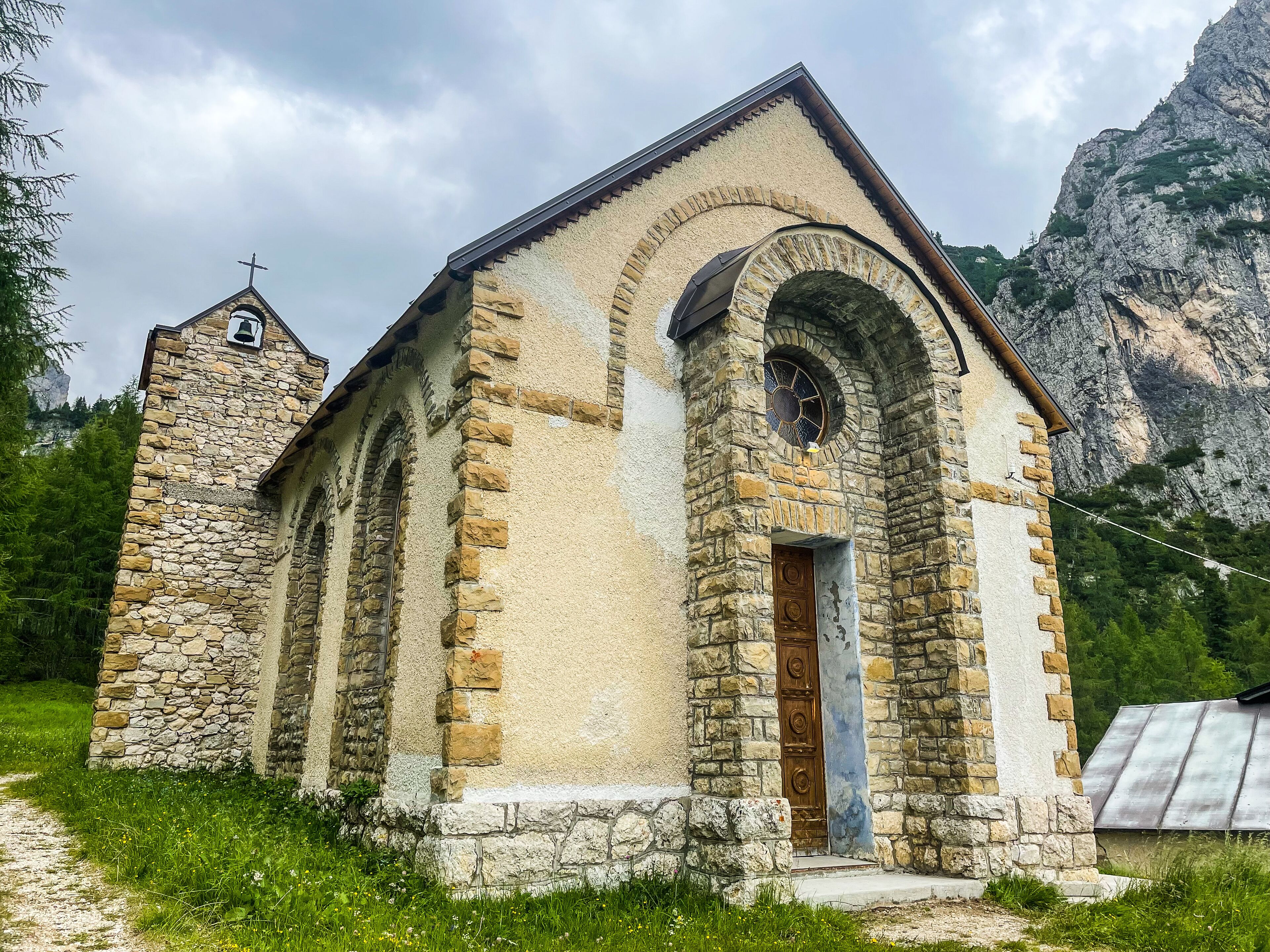 Una pequeña iglesia de piedra con torre campanario y entrada abovedada, rodeada de follaje y montañas.