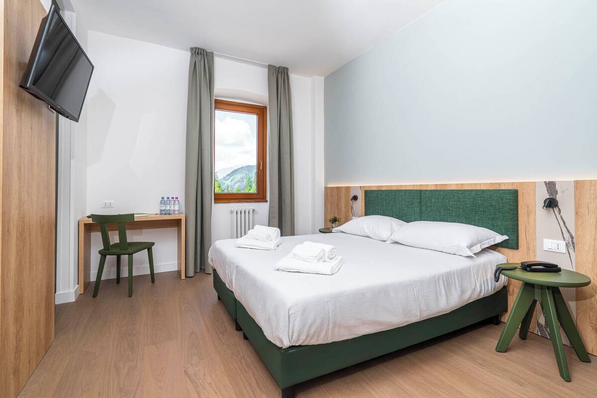 Une chambre d’hôtel équipée d’un lit, d’un bureau, d’une chaise, d’une télévision et d’une fenêtre donnant sur l’extérieur.