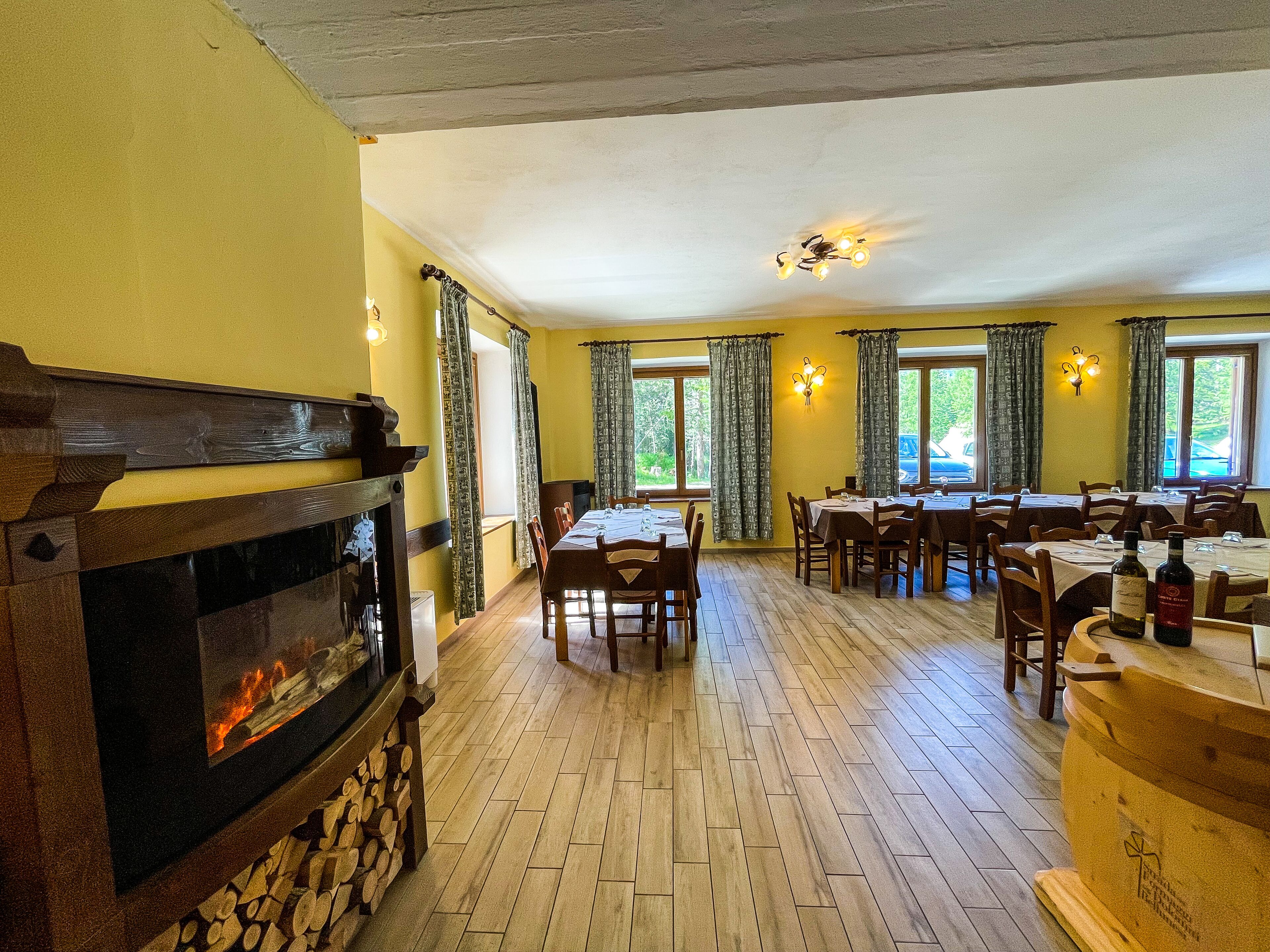 A dining area with wooden tables and chairs, a fireplace, and large windows with curtains.