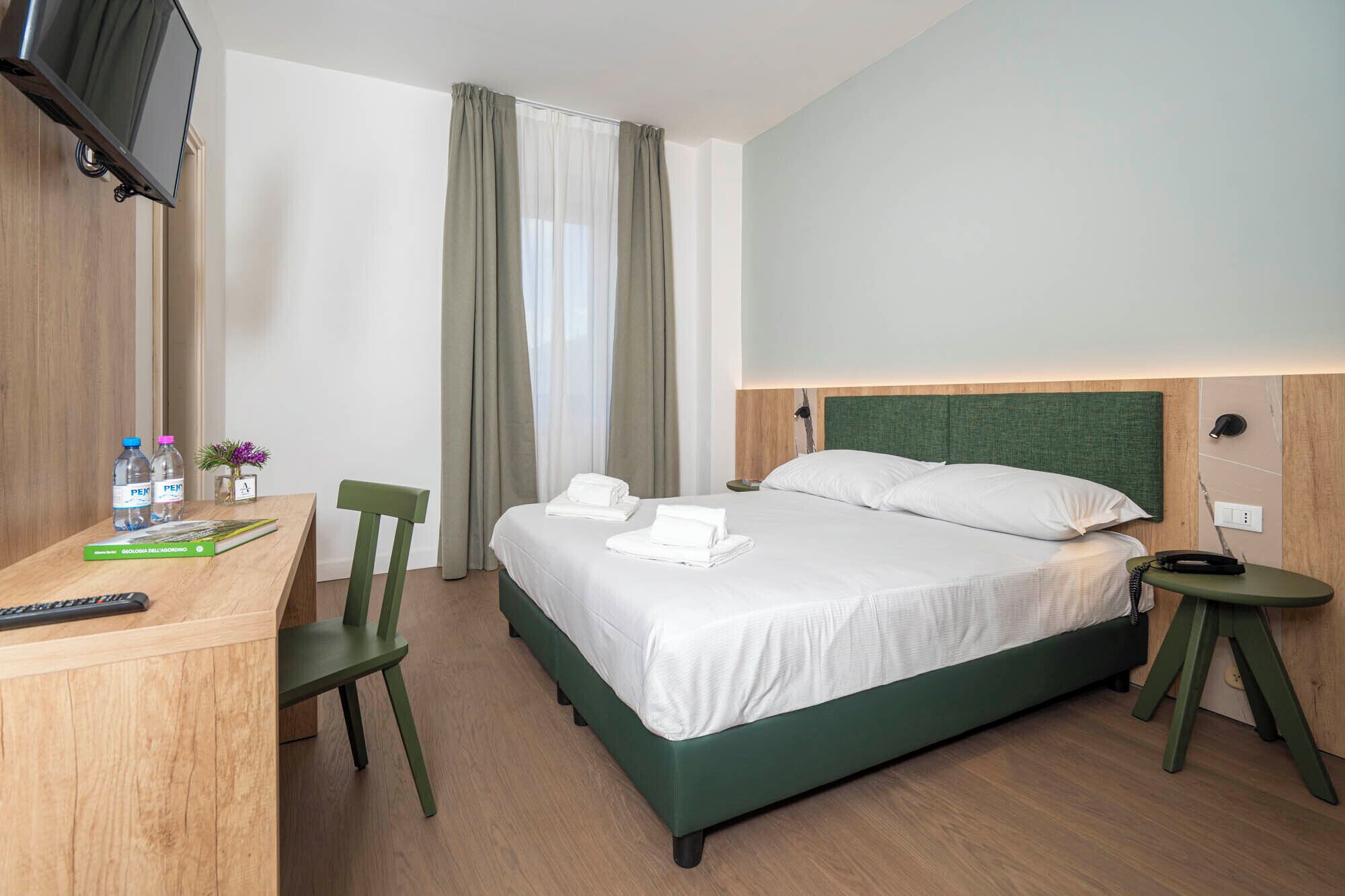 A hotel room with a bed, a desk with a chair, a TV mounted on the wall, and a window with curtains.