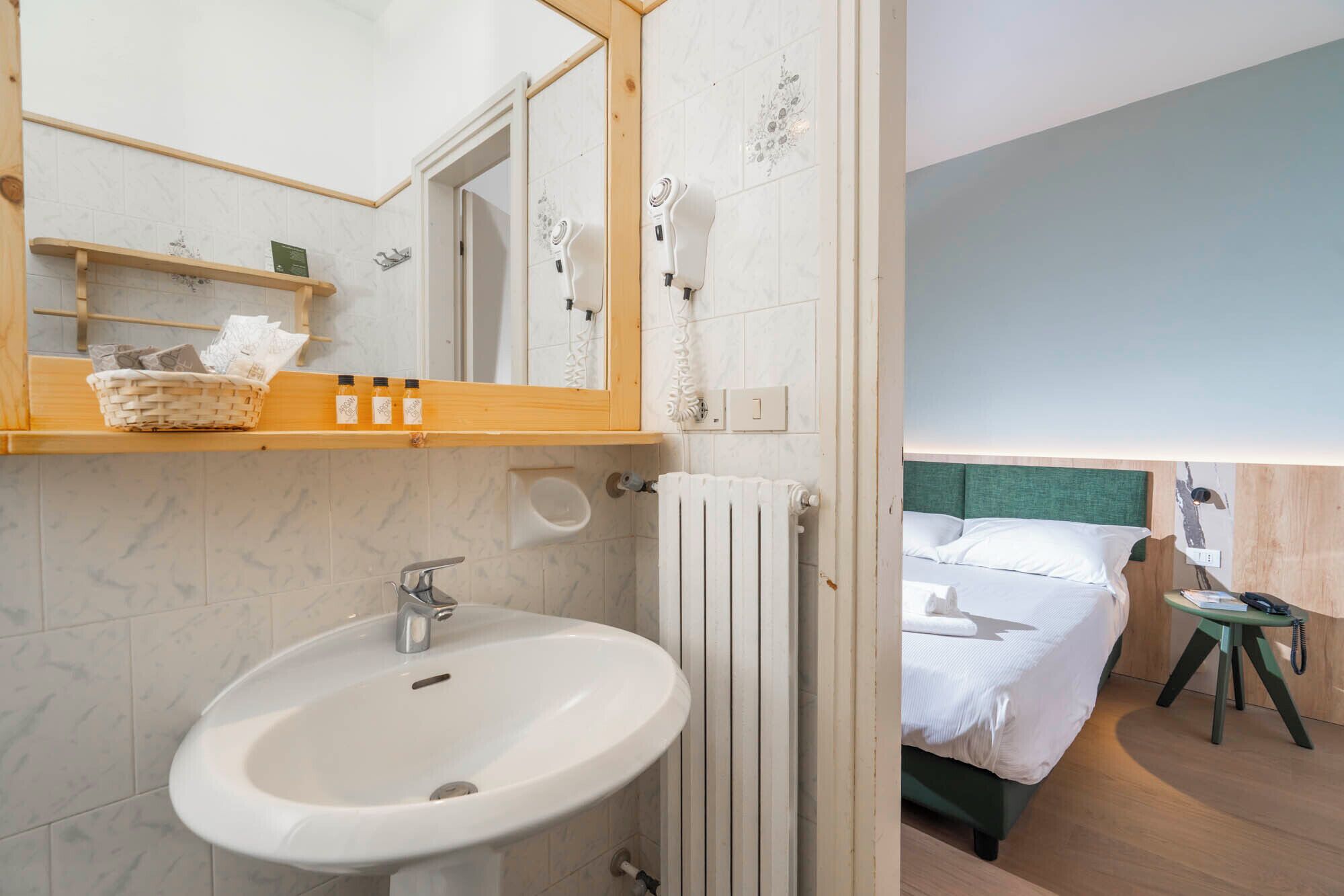 Ein Badezimmer mit einem weißen Waschbecken, einem Spiegel und einem Handtuchhalter. Es gibt ein Holzregal mit einem Korb und Toilettenartikeln. Neben dem Bett ist eine Heizung sichtbar.