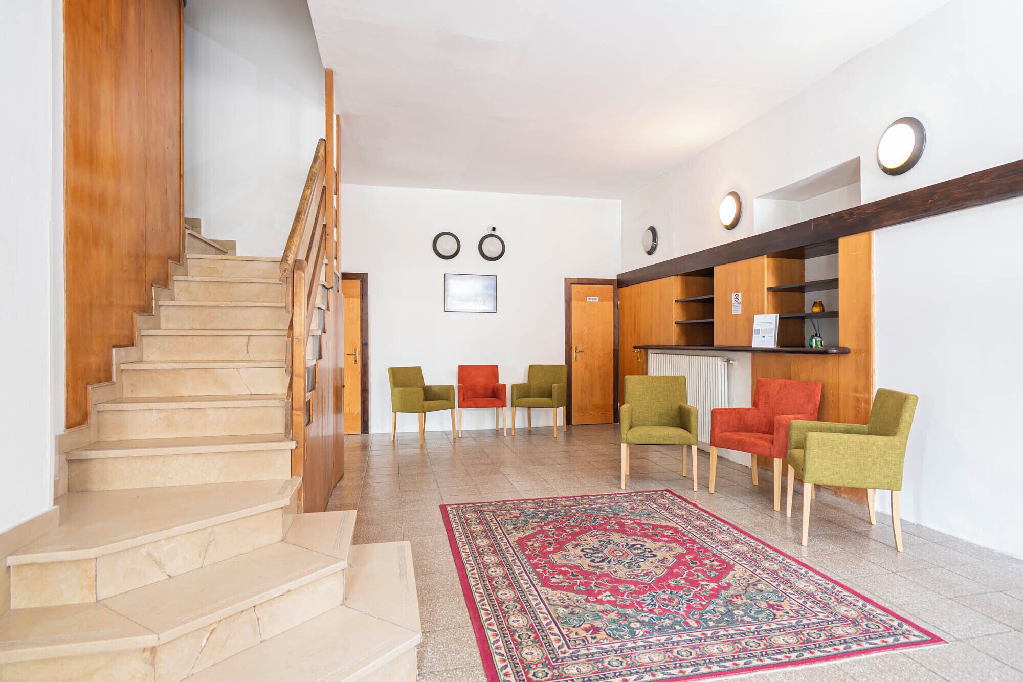 Una escalera conduce a una sala de estar con un tapete rojo estampado, estantes de madera y varias sillas.