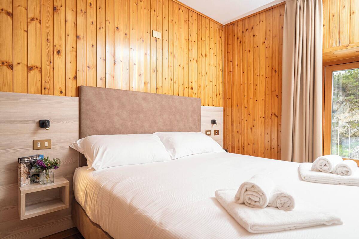 Une chambre d’hôtel avec un mur lambrissé en bois, un lit avec du linge de maison blanc, une table de chevet avec une lampe et une fenêtre avec des rideaux.