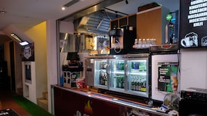Zona de bar con un cartel de Shiraz, un refrigerador lleno de bebidas y una barra con publicidad de Shiraz.