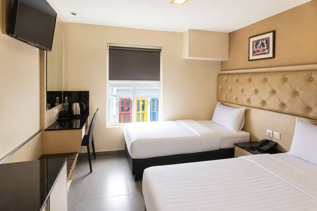 A hotel room with two beds, a TV, a desk, a chair, a window with a view of colorful buildings, and a framed picture on the wall.