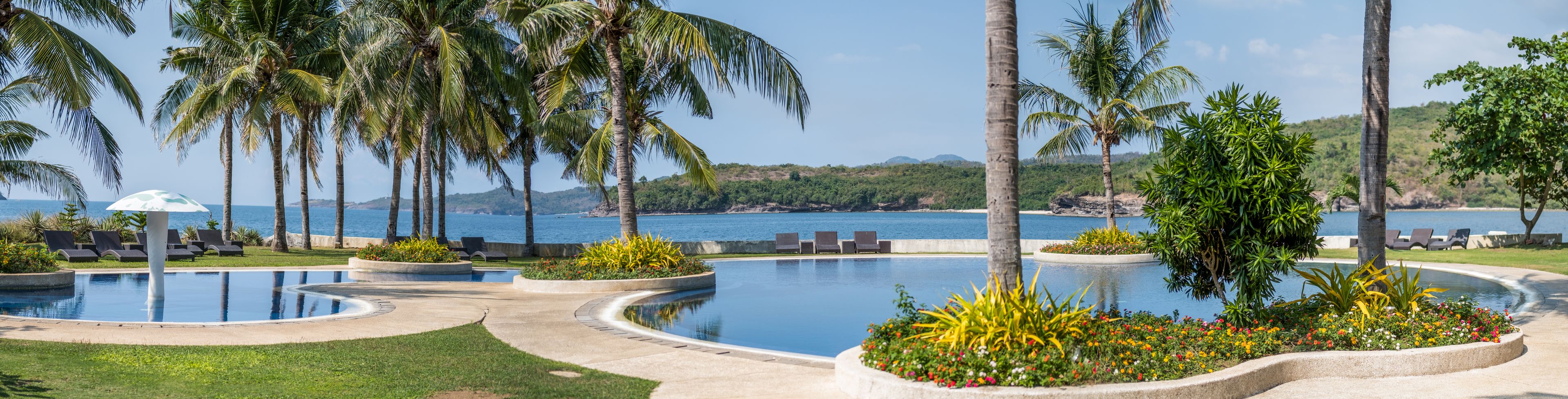 A resort pool area with multiple swimming pools, palm trees, and a view of the ocean.