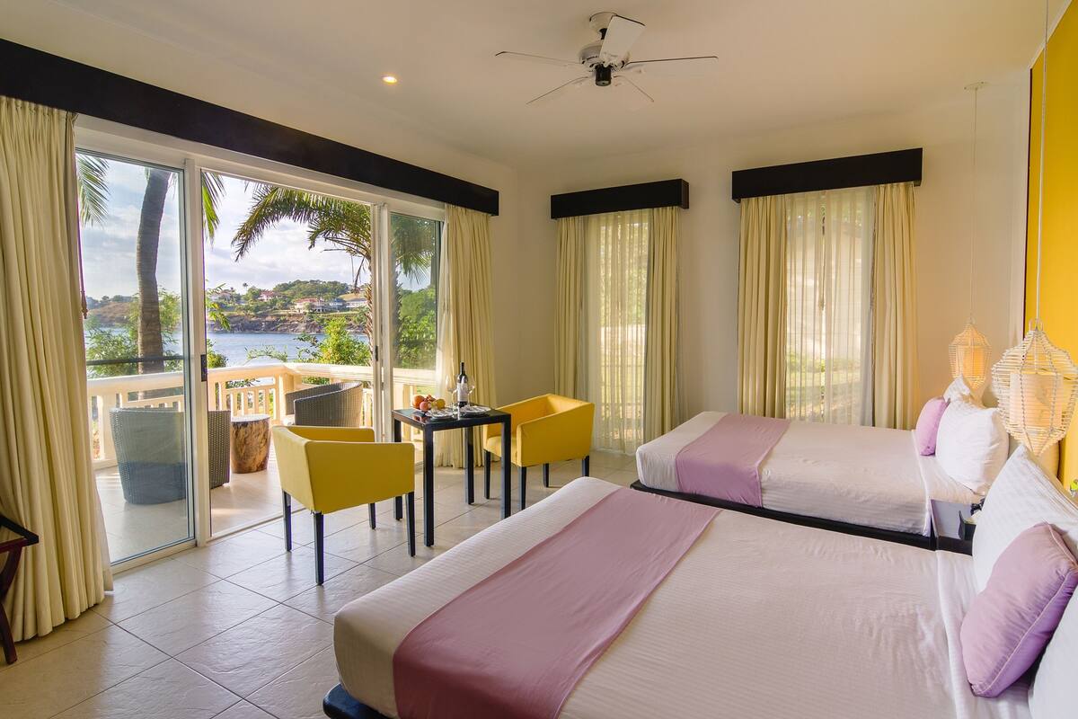 A hotel room with two beds, a balcony with yellow chairs, a table with a bottle, and a view of the sea.