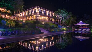 A large, illuminated hotel with a reflection in the water, a bridge, and a pool area with umbrellas. - Club Punta Fuego (Nasugbu)