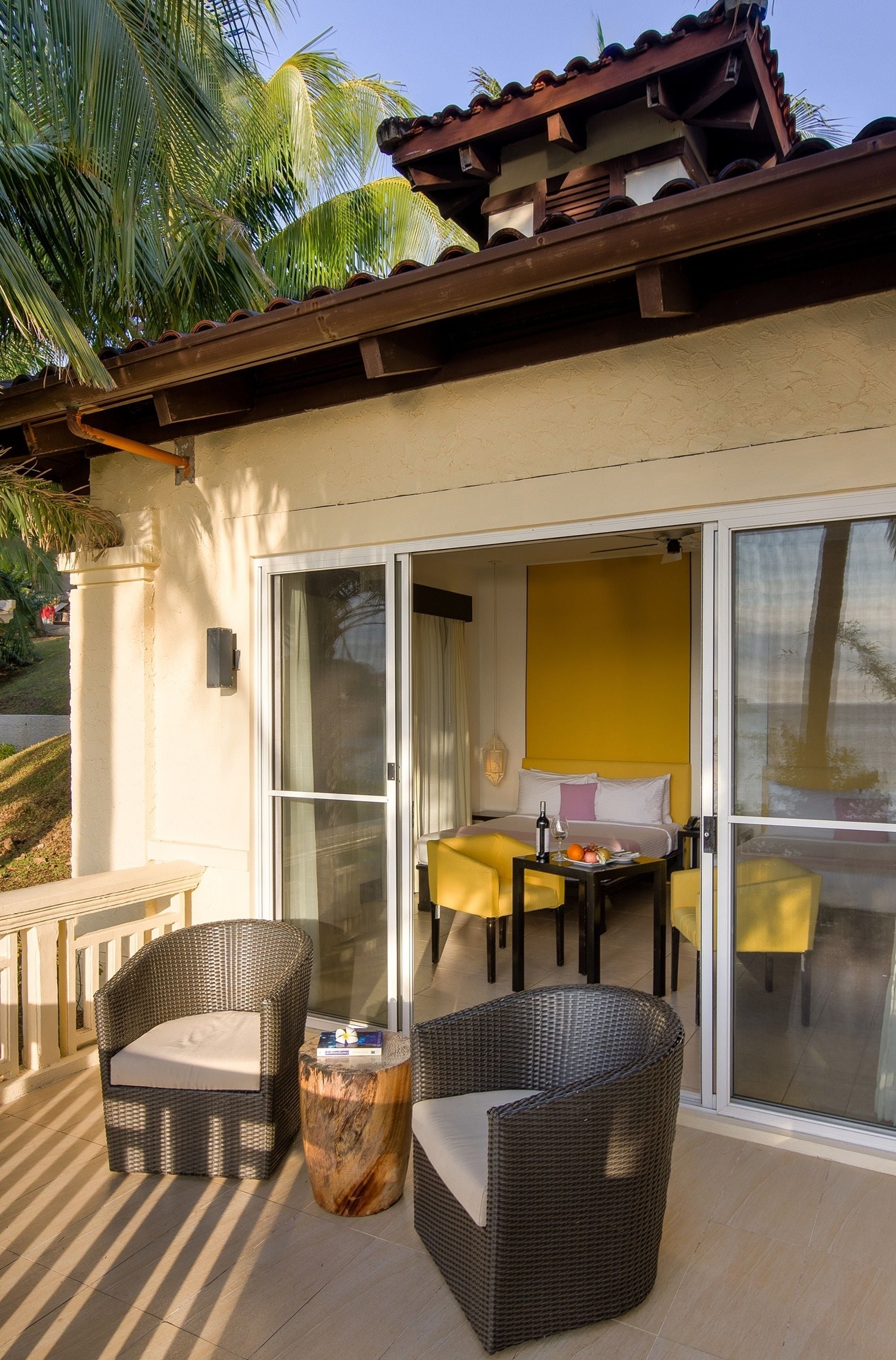 Un balcon avec des meubles en osier, une table garnie de fruits et un mur jaune à l’intérieur.