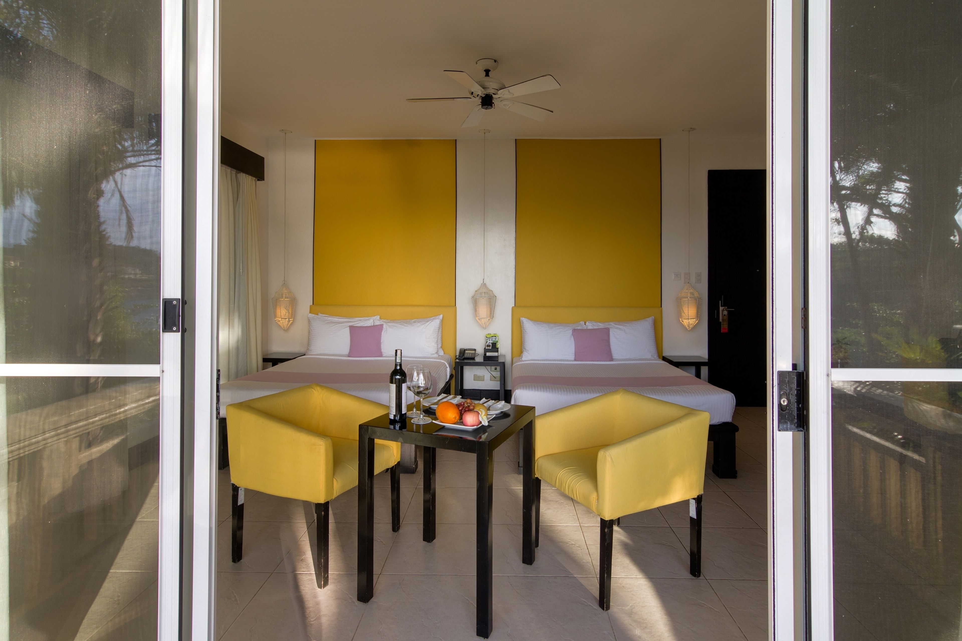 A room with a bed, two yellow chairs, a table with a bottle and fruit, and a ceiling fan.