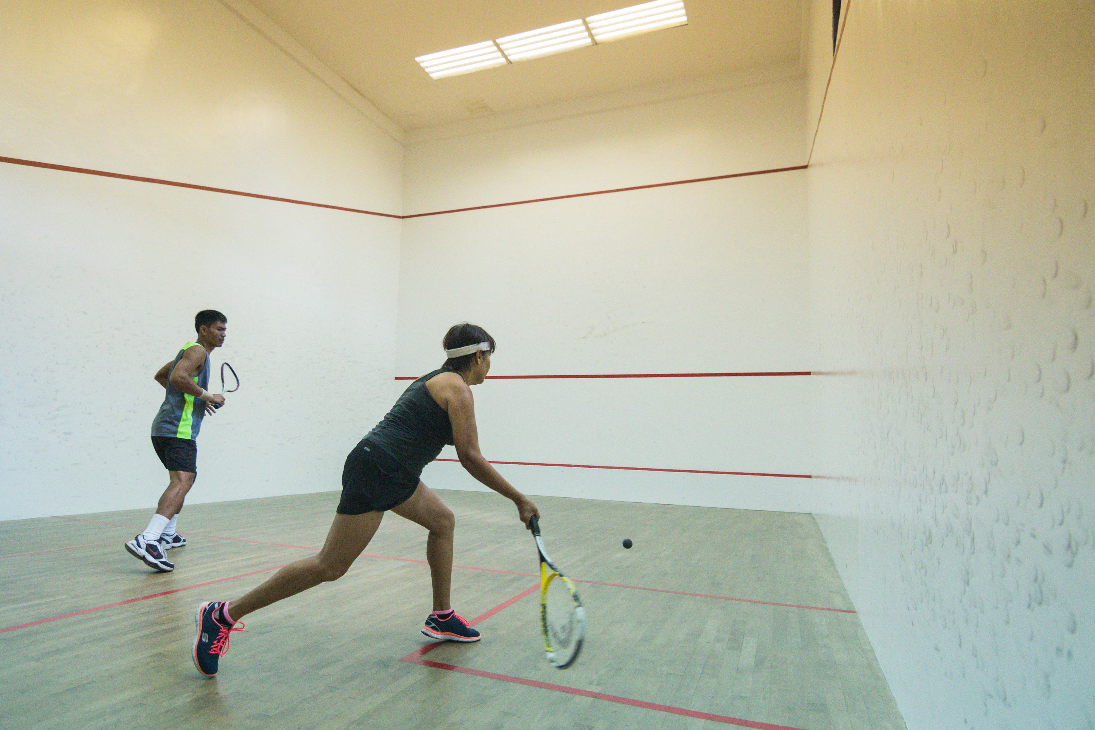 Una cancha de squash con dos jugadores, uno vestido de negro y el otro con un atuendo gris y verde, jugando activamente.