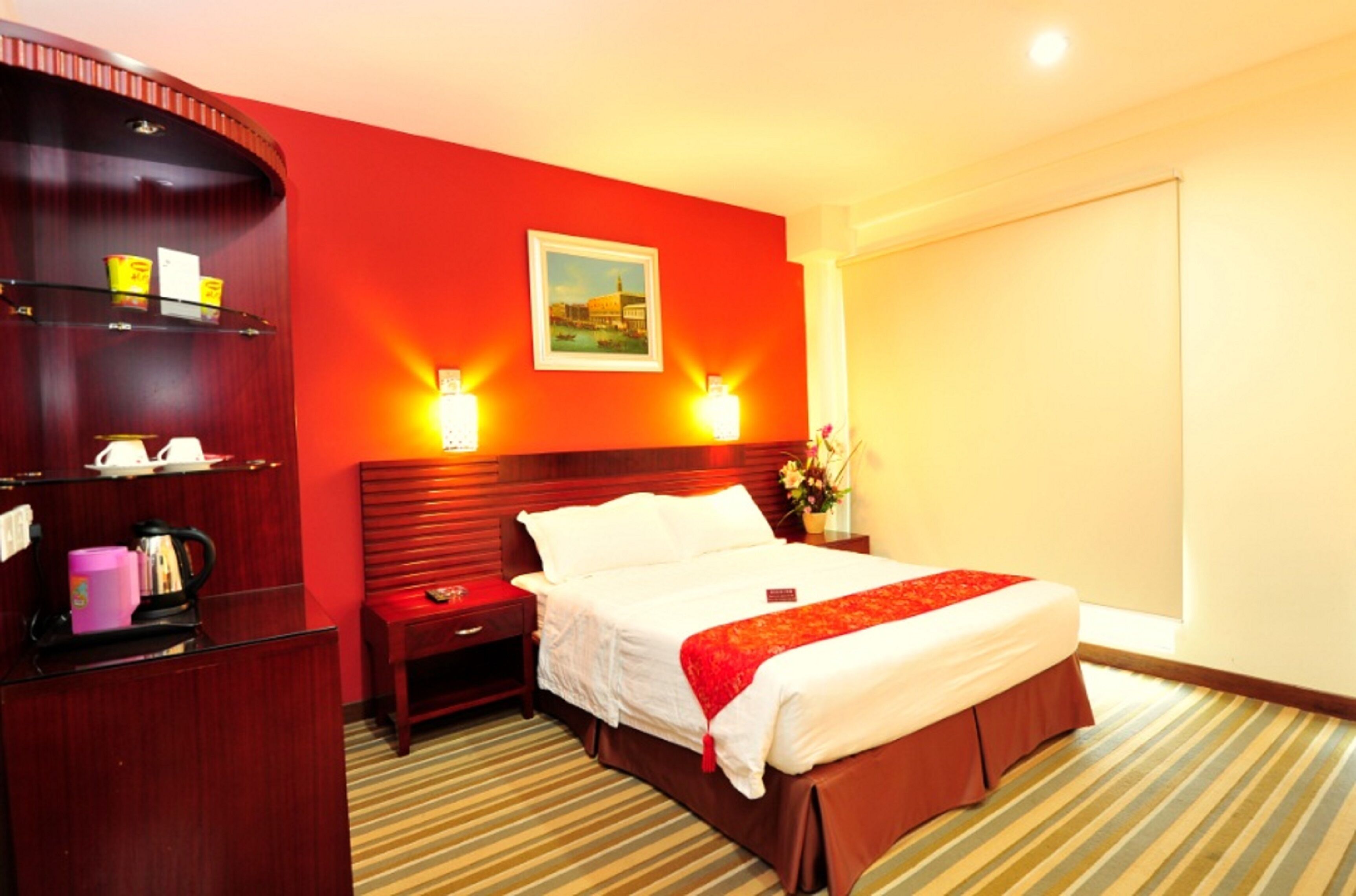 A hotel room with a large bed, a wooden nightstand, a red wall, and a painting above the bed.