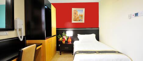 A hotel room with a bed, a desk, a chair, a nightstand with a lamp, and a painting on the wall.