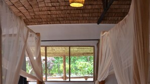 A room with two wooden beds, a woven ceiling, a large window with a view of greenery, and a wicker chair. - Honey Badger Lodge (Moshi)