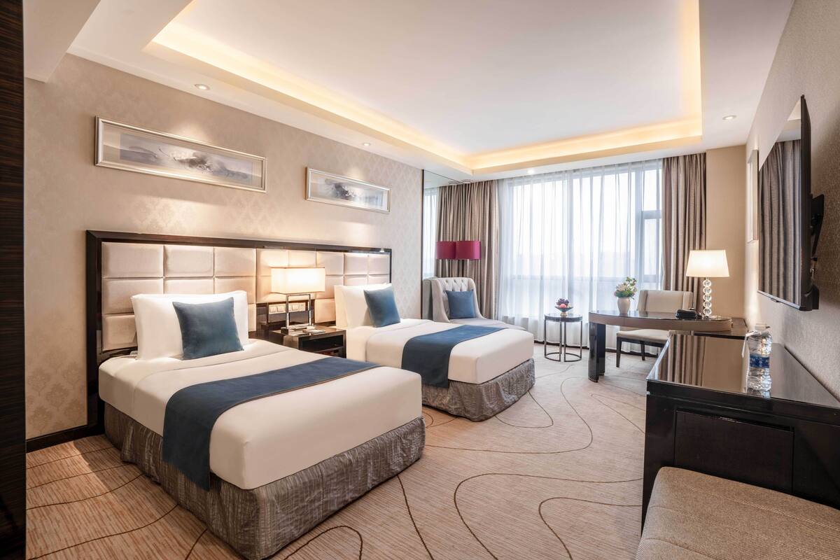 Habitación de hotel con dos camas, un escritorio, una silla, un televisor y un ventanal con cortinas.