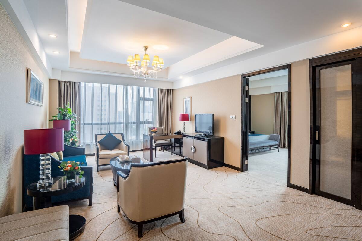 Habitación de hotel con zona de estar, televisión, escritorio y ventana grande con cortinas.