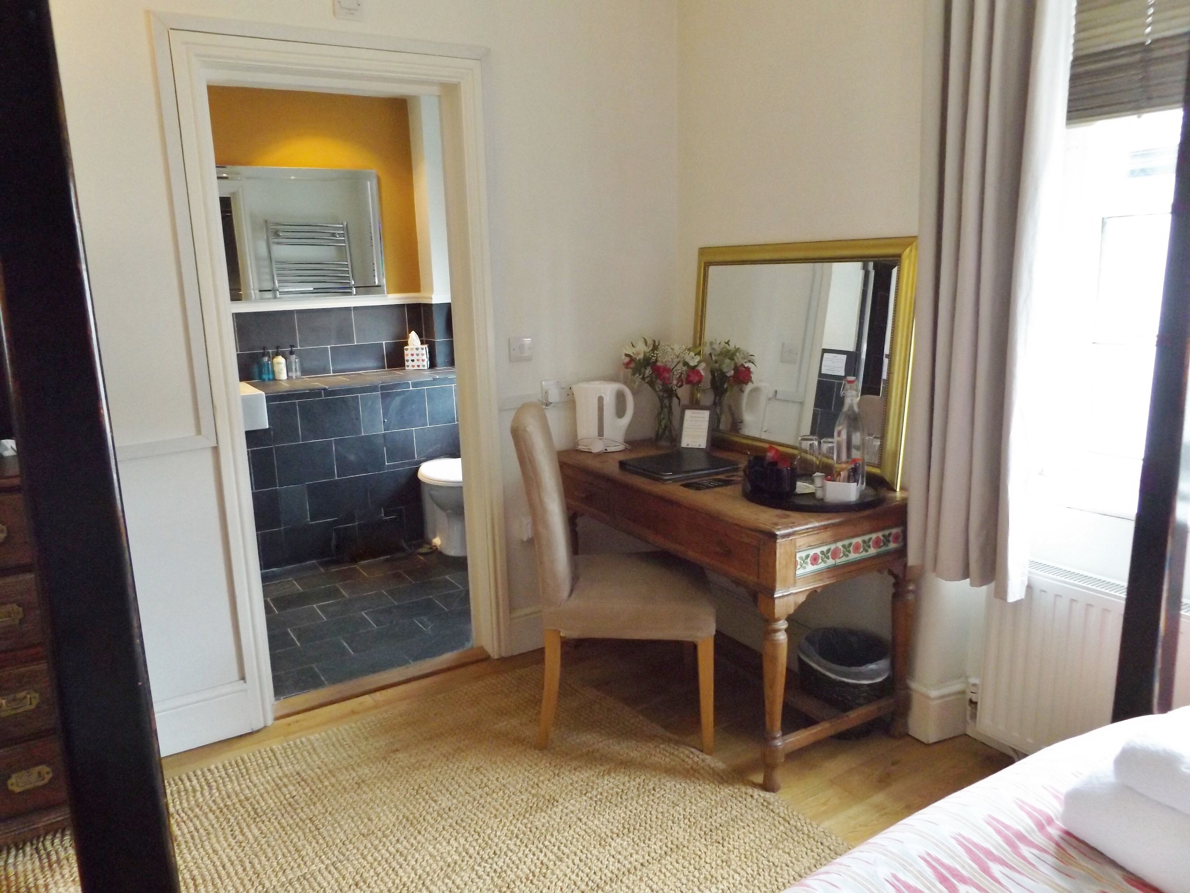 Habitación de hotel con escritorio de madera, espejo, silla, cama, baño con lavamanos e inodoro, y ventana con cortinas.