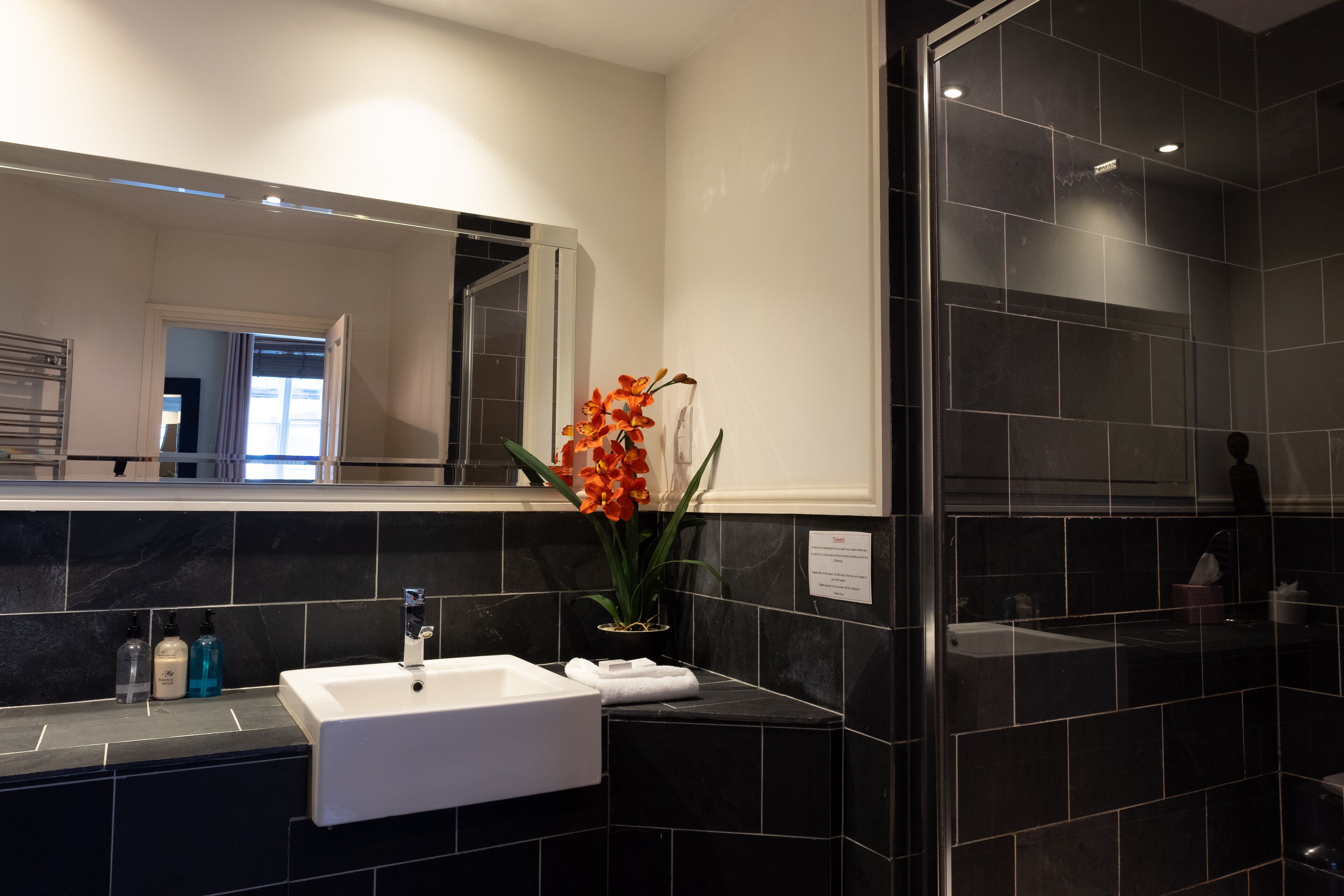 A modern bathroom with a white sink, black tiles, a glass shower enclosure, and a potted orange orchid.