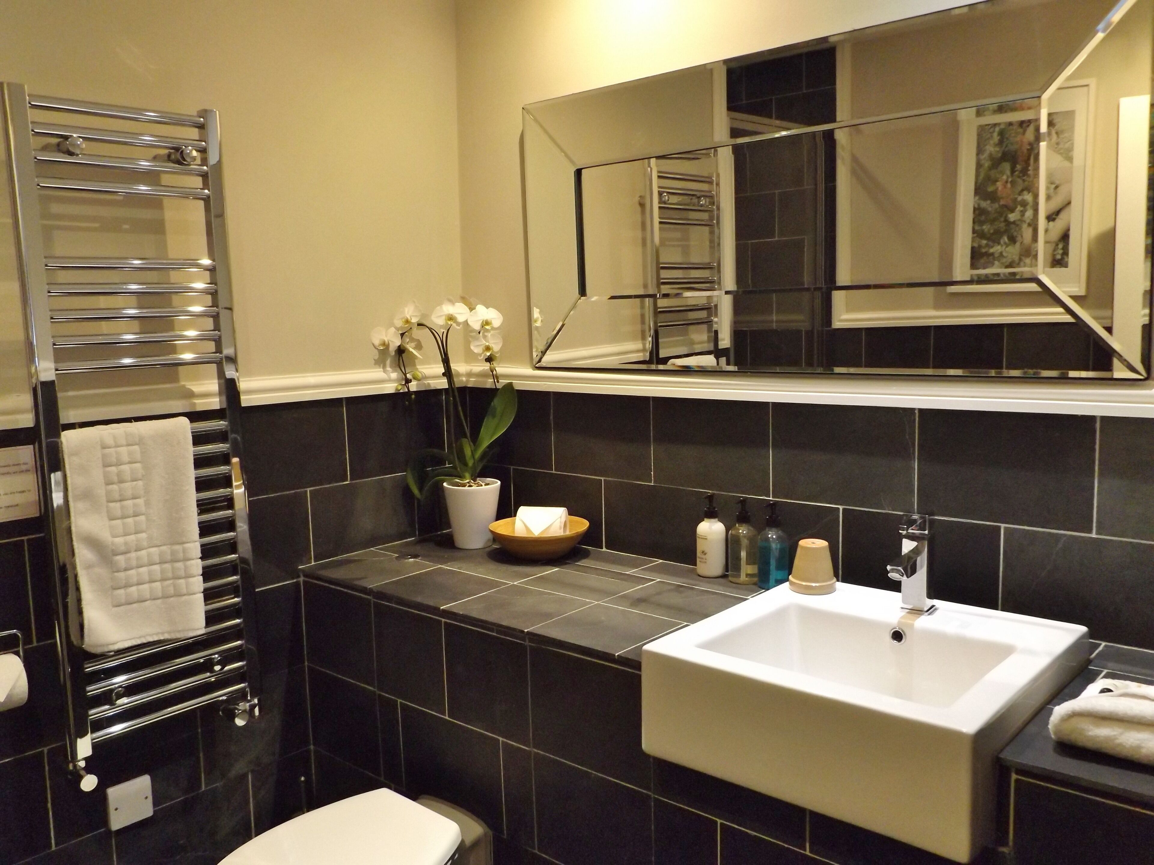A modern bathroom with a white sink, black tiles, a glass shower enclosure, a towel rack, and a potted orchid.