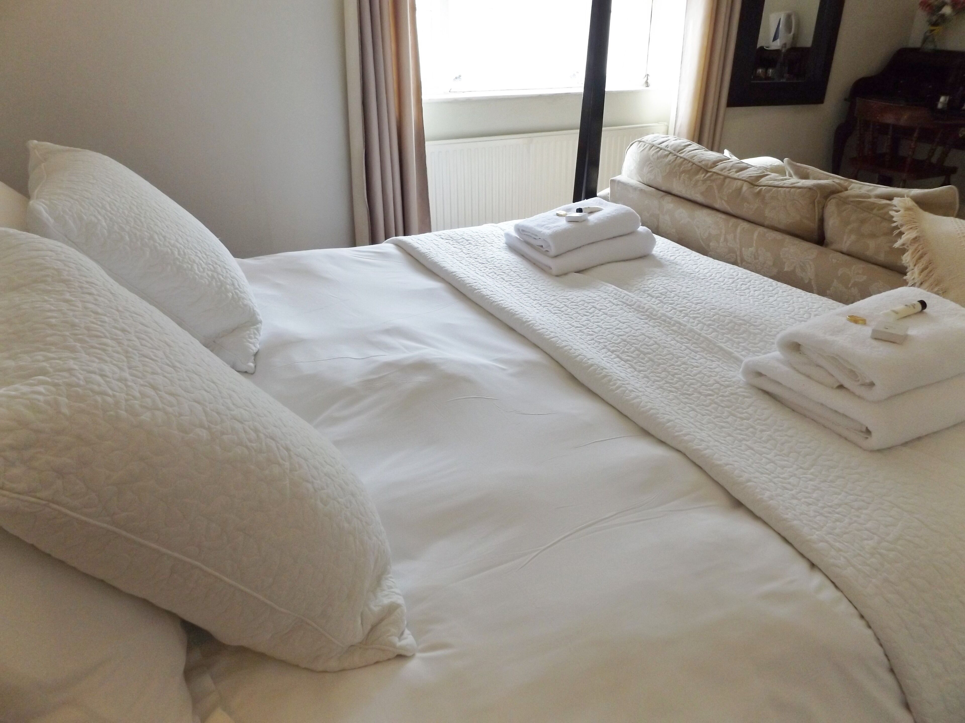 Una cama bien hecha con sábanas y almohadones blancos, una toalla doblada sobre la cama y una ventana con cortinas.