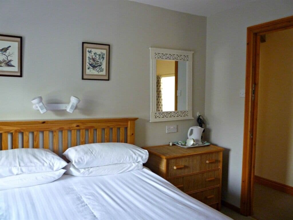 A hotel room with a wooden headboard, a bed with white linens, a bedside table with a telephone and a cup, and two framed pictures on the wall.