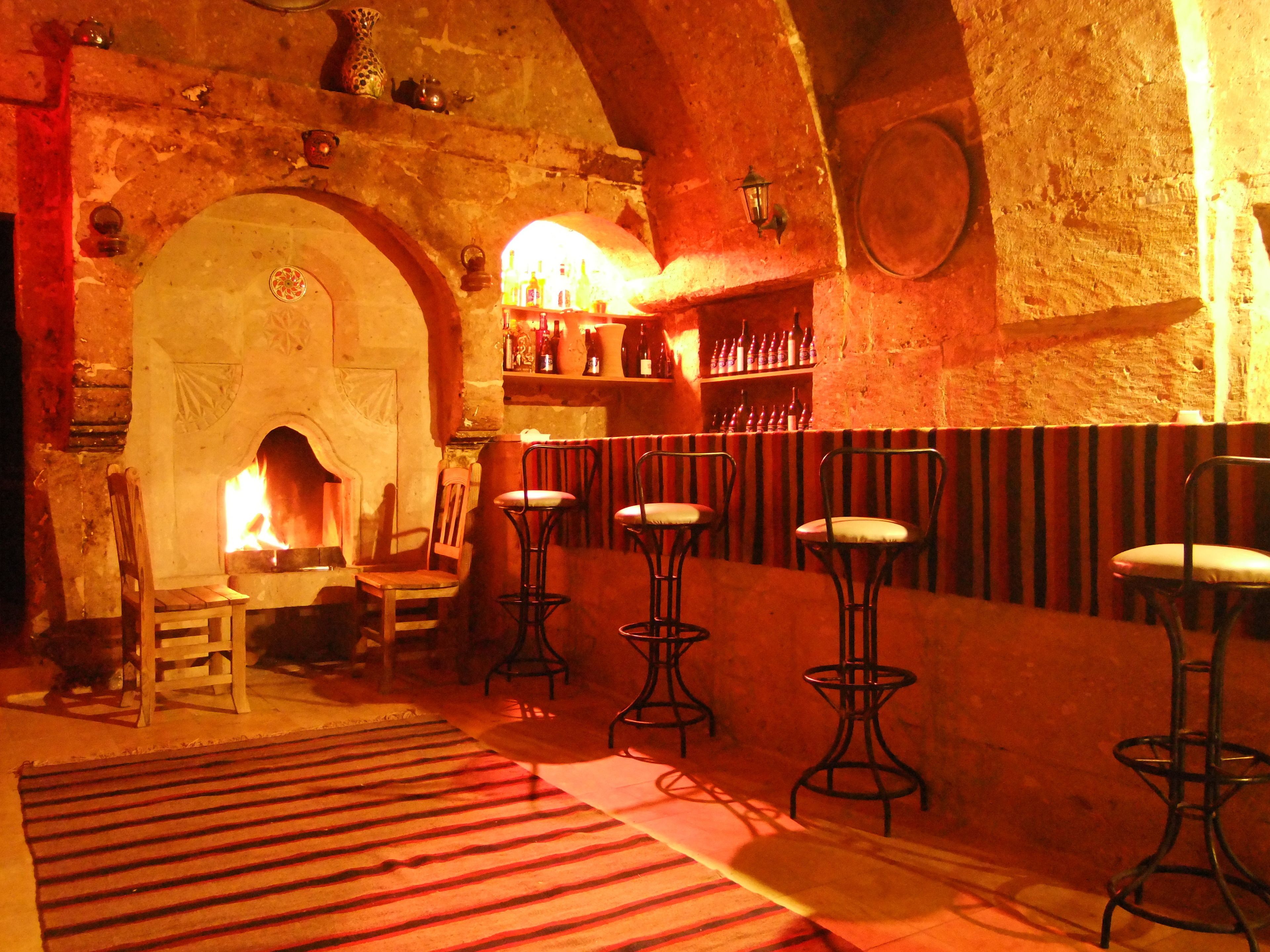 A rustic interior with a stone fireplace, wooden chairs, and a bar area with bottles.