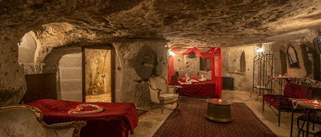 Ein Zimmer mit rustikalem Steinbezug an der Decke, einem roten Teppich und einem Bett mit rotem Baldachin. Zwei Stühle und ein Tisch mit Kerzen sind ebenfalls vorhanden.