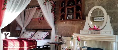 Un dormitorio con cama de dosel, un aparador de madera con objetos decorativos, un tocador con espejo y dos copas de vino sobre un mantel rojo.