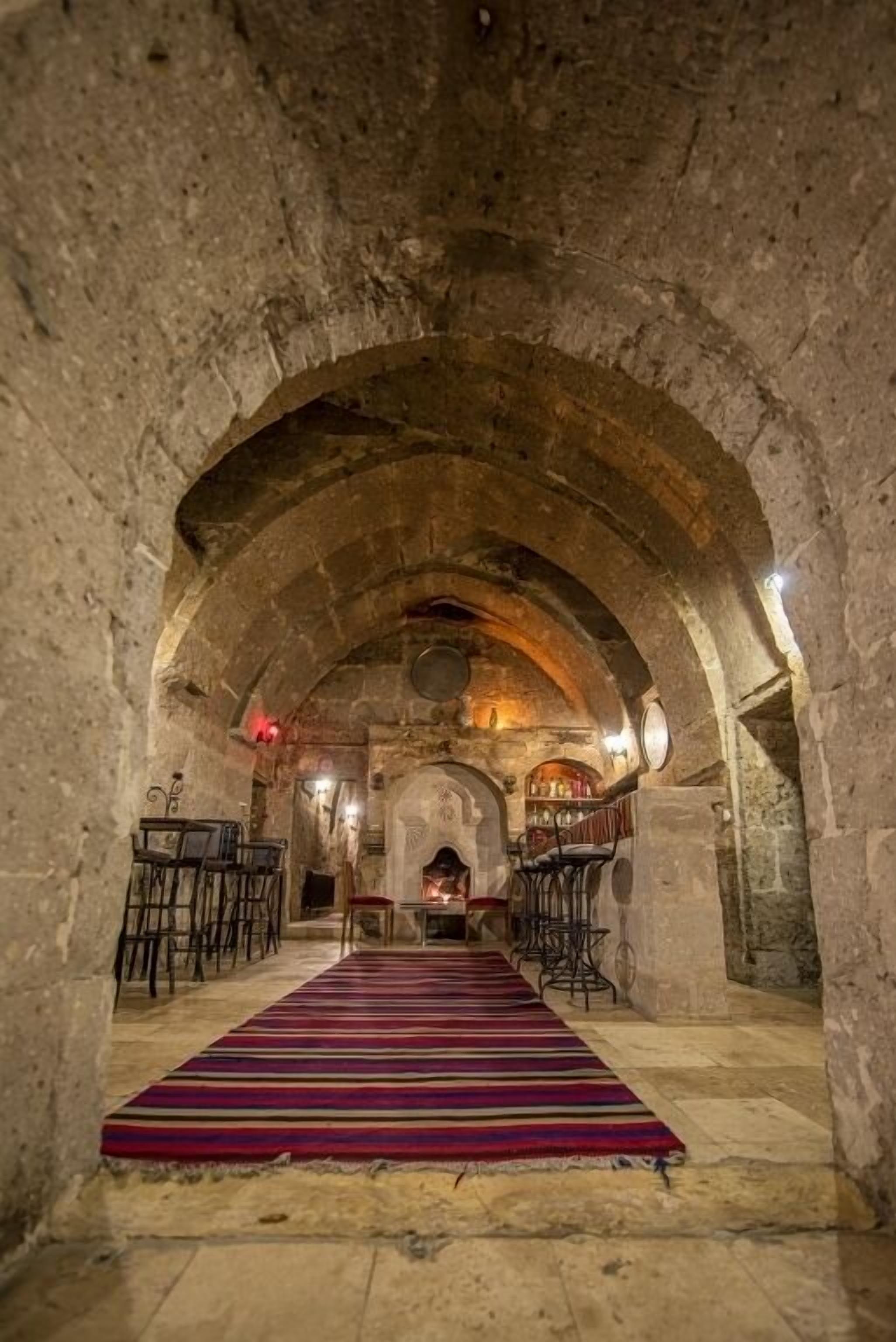 An arched stone corridor with a patterned carpet, tables, and chairs, leading to a central area with a fireplace.