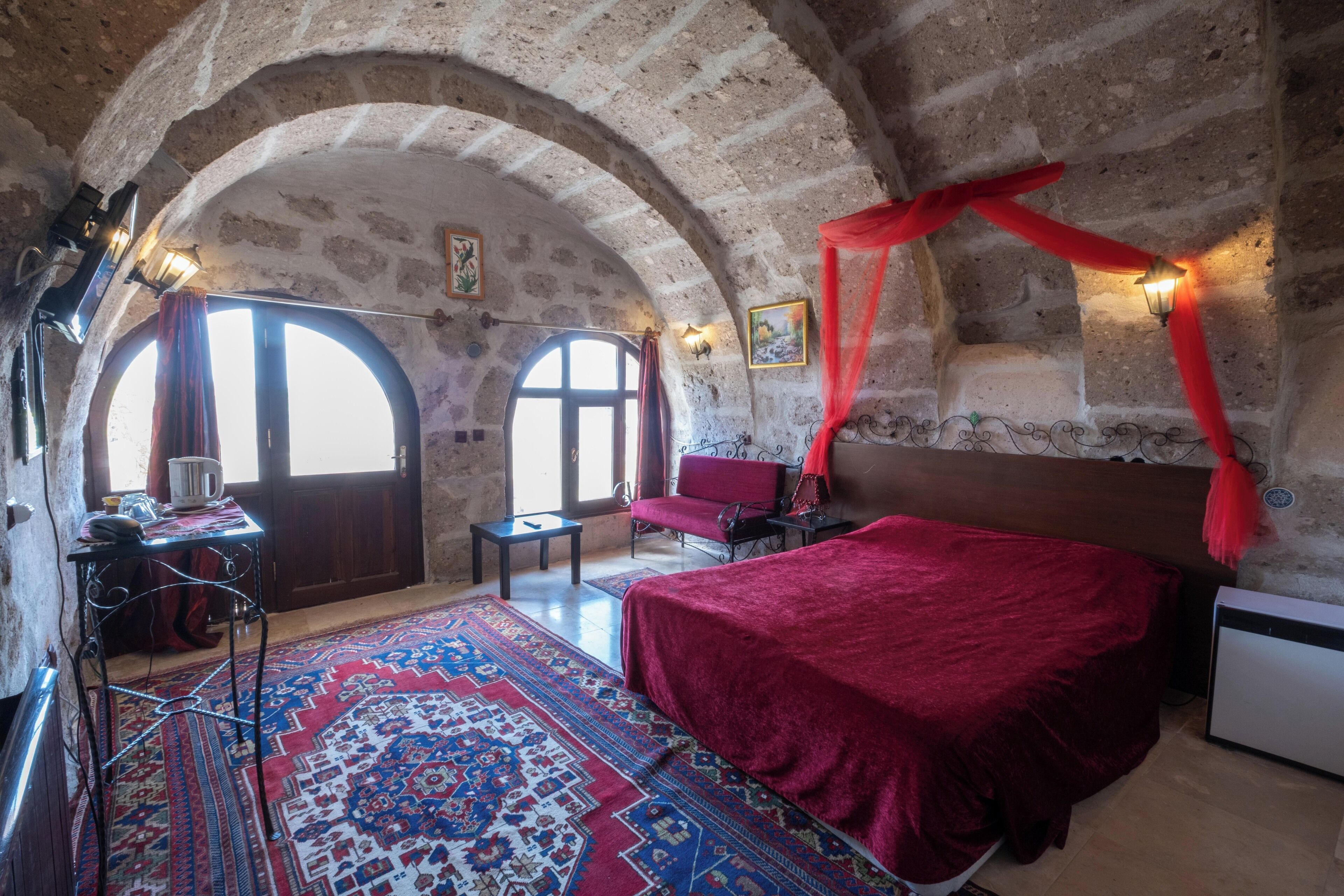 Una habitación con una cama grande, un escritorio y una silla. Hay dos ventanas con cortinas, un dosel rojo sobre la cama y una alfombra estampada en el suelo.