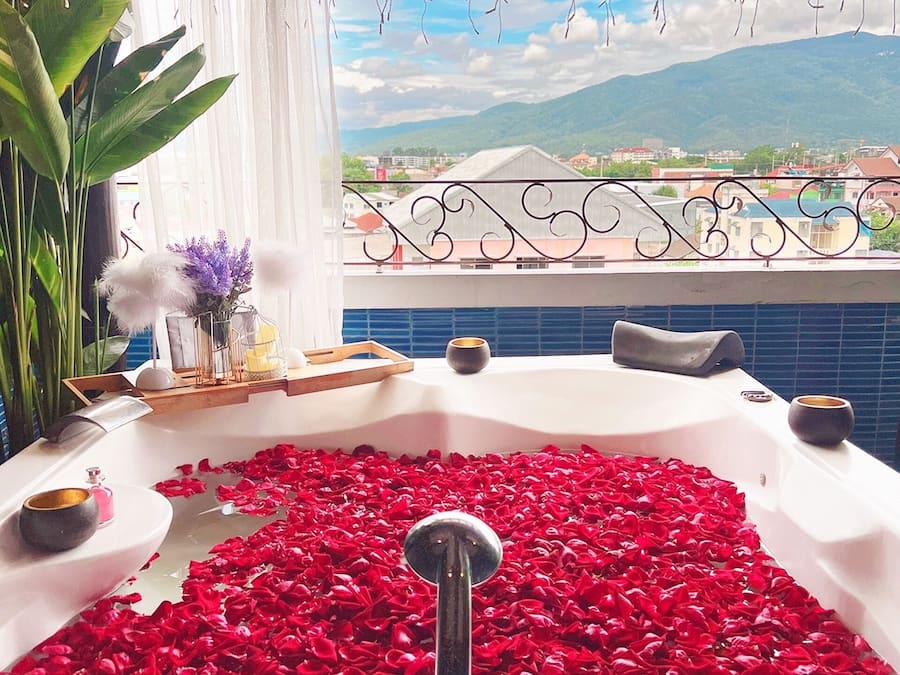 Uma banheira com pétalas de rosa, rodeada por objetos decorativos e vista para uma paisagem montanhosa.
