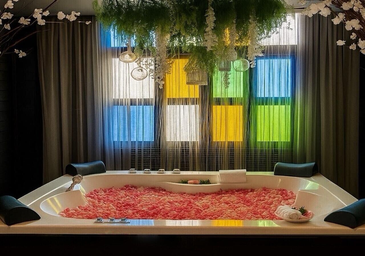 Een spa-ruimte met een groot bad vol rozenblaadjes, omringd door donkere gordijnen en uitzicht op kleurrijke gebouwen.