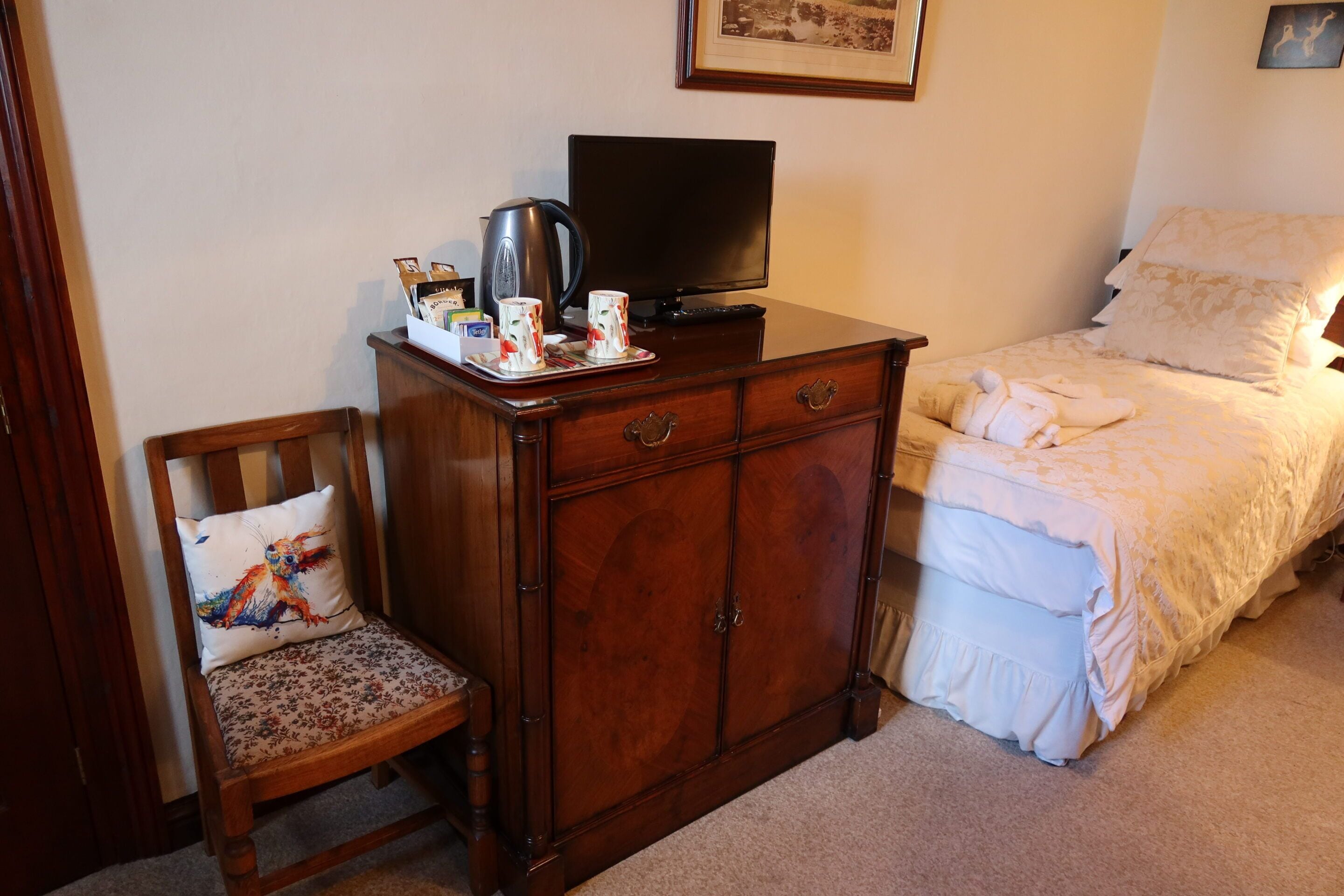 Ein Schlafzimmer mit einem hölzernen Schminktisch, einem Fernseher, einem Bett mit weißer Bettwäsche und einem Sessel mit einem blumigen Kissen.