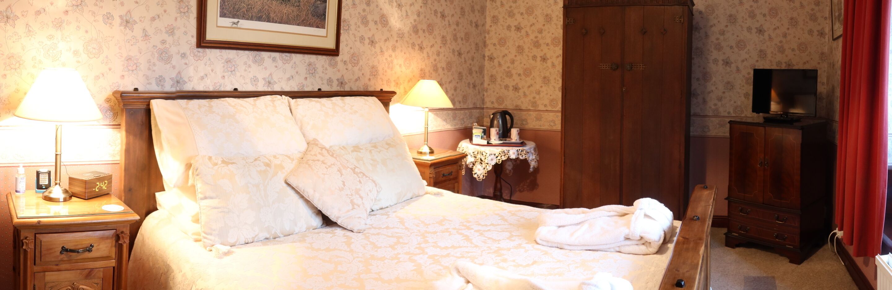 Un lit bien fait, avec une literie à motifs floraux, une table de chevet avec une lampe, une armoire en bois et un téléviseur posé sur un meuble.