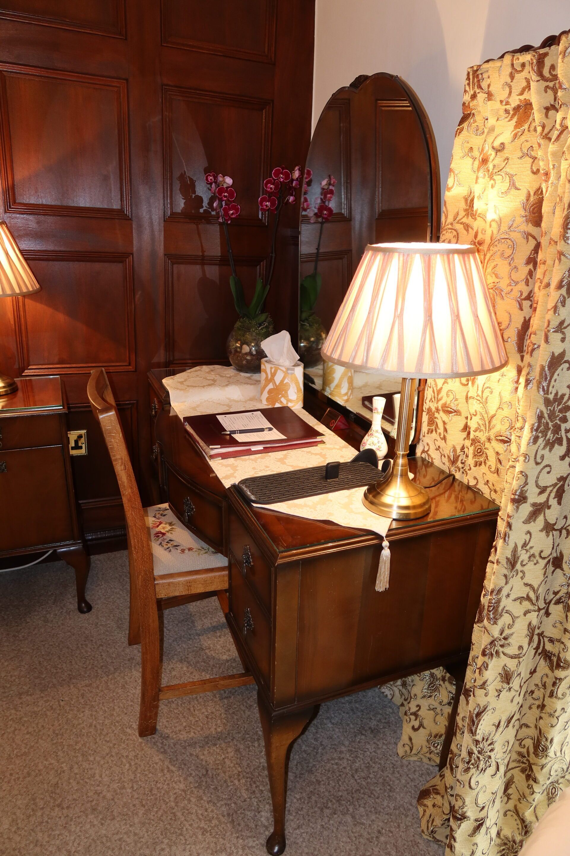 A wooden desk with a lamp, a chair, and a vase with flowers.