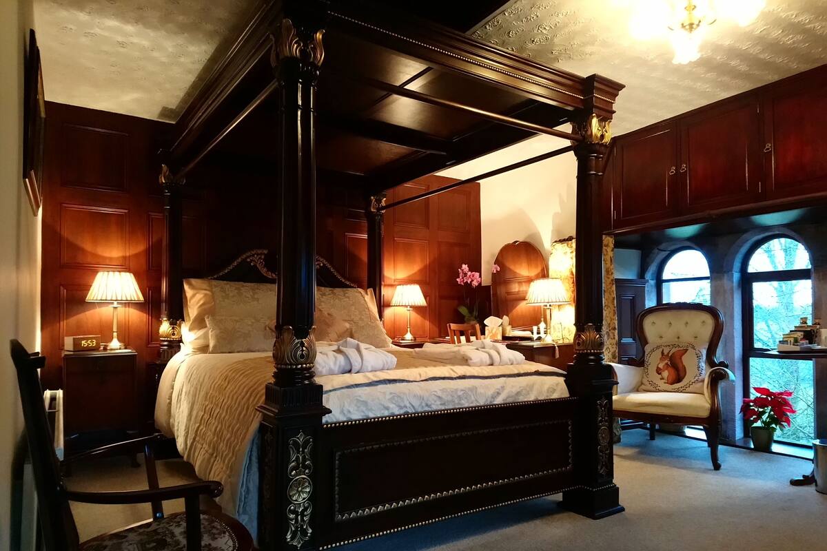 A four-poster bed with a canopy, a sitting chair, a desk with a lamp, and a view of the outdoors through arched windows.