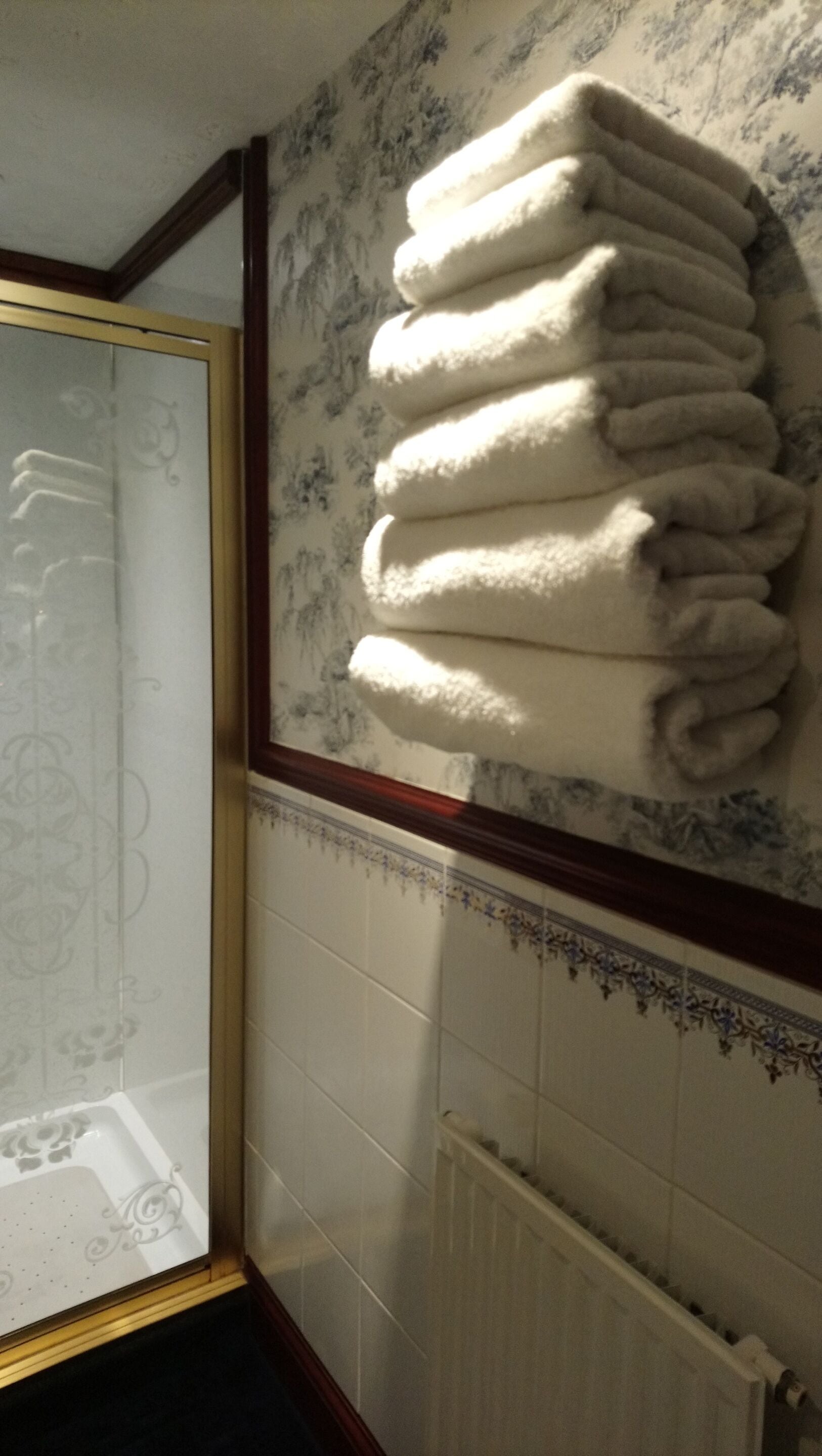 A bathroom with a stack of white towels on a wall, a glass shower door, and a radiator.