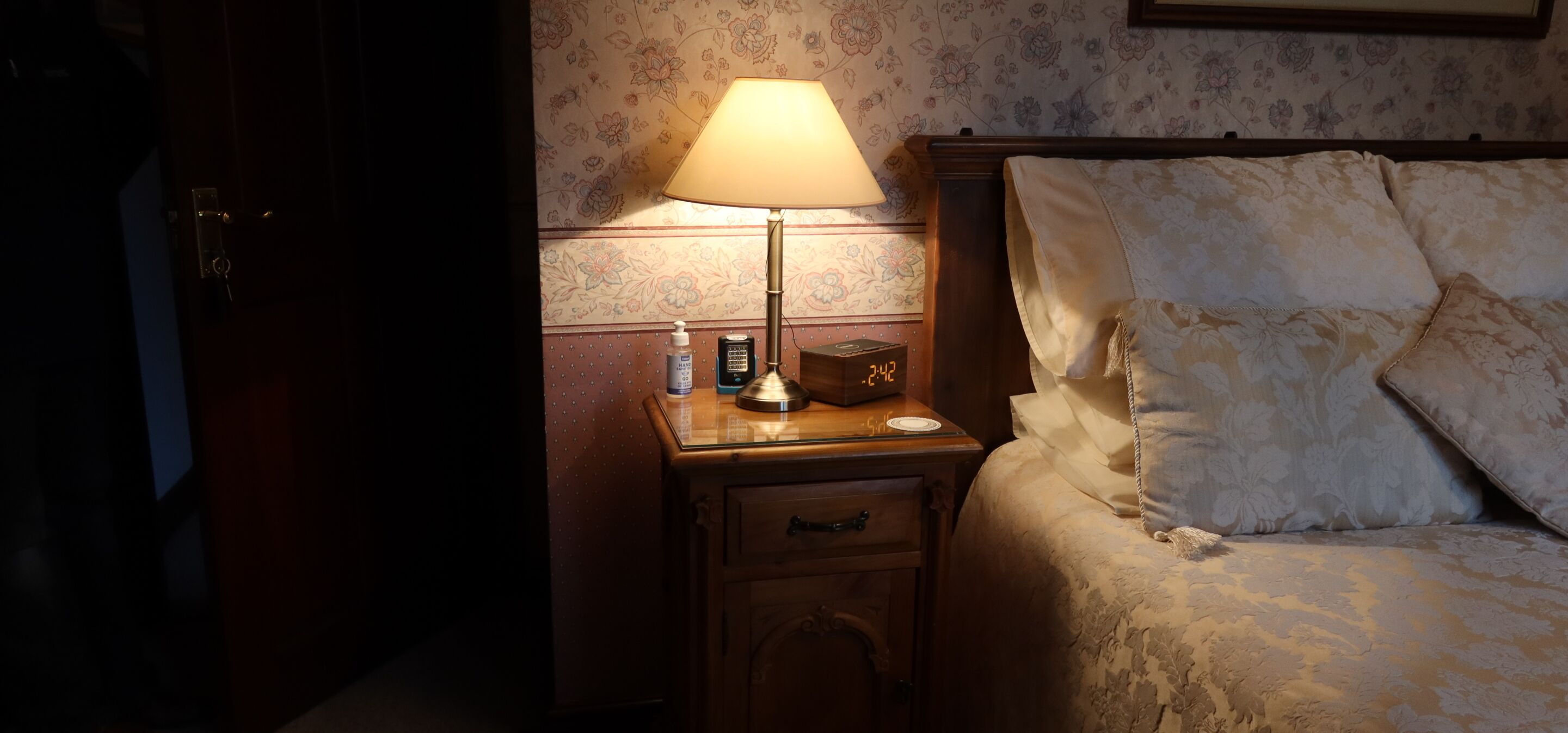 A hotel room with a bed, a nightstand with a lamp, a clock, and a small table with a bottle and a box.