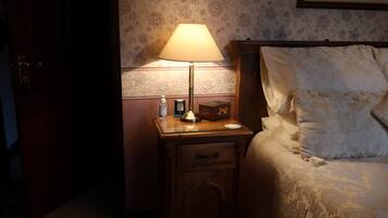 Une chambre d’hôtel avec un lit, une table de chevet avec une lampe, une horloge et une petite table sur laquelle se trouvent une bouteille et une boîte.