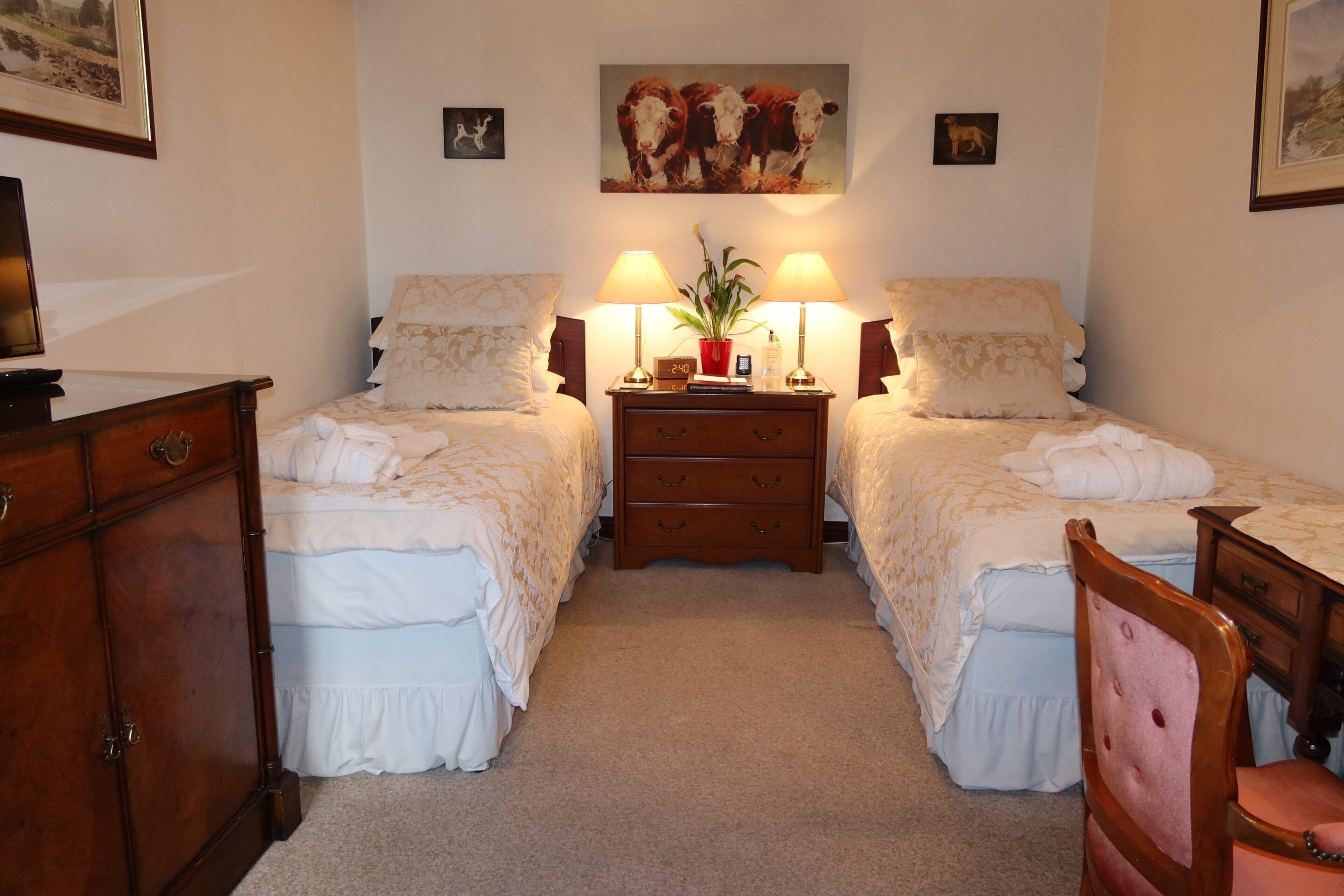 A bedroom with two beds, a dresser, a nightstand with lamps, and a painting of cows on the wall.