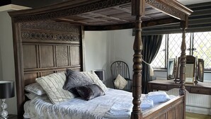 Una cama con dosel de madera oscura con intrincados grabados, una mesita de noche con una lámpara y una ventana con vistas a árboles.