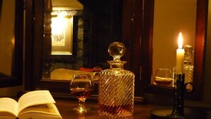 Una mesa de madera con un libro, una copa de vino tinto, un decantador de cristal y una vela encendida.