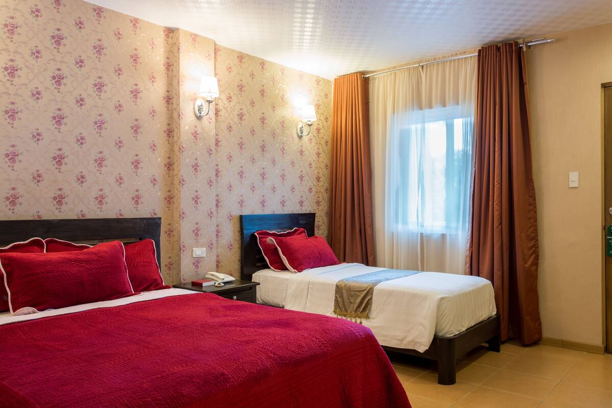 Quarto de hotel com duas camas, papel de parede floral e uma janela com cortinas.