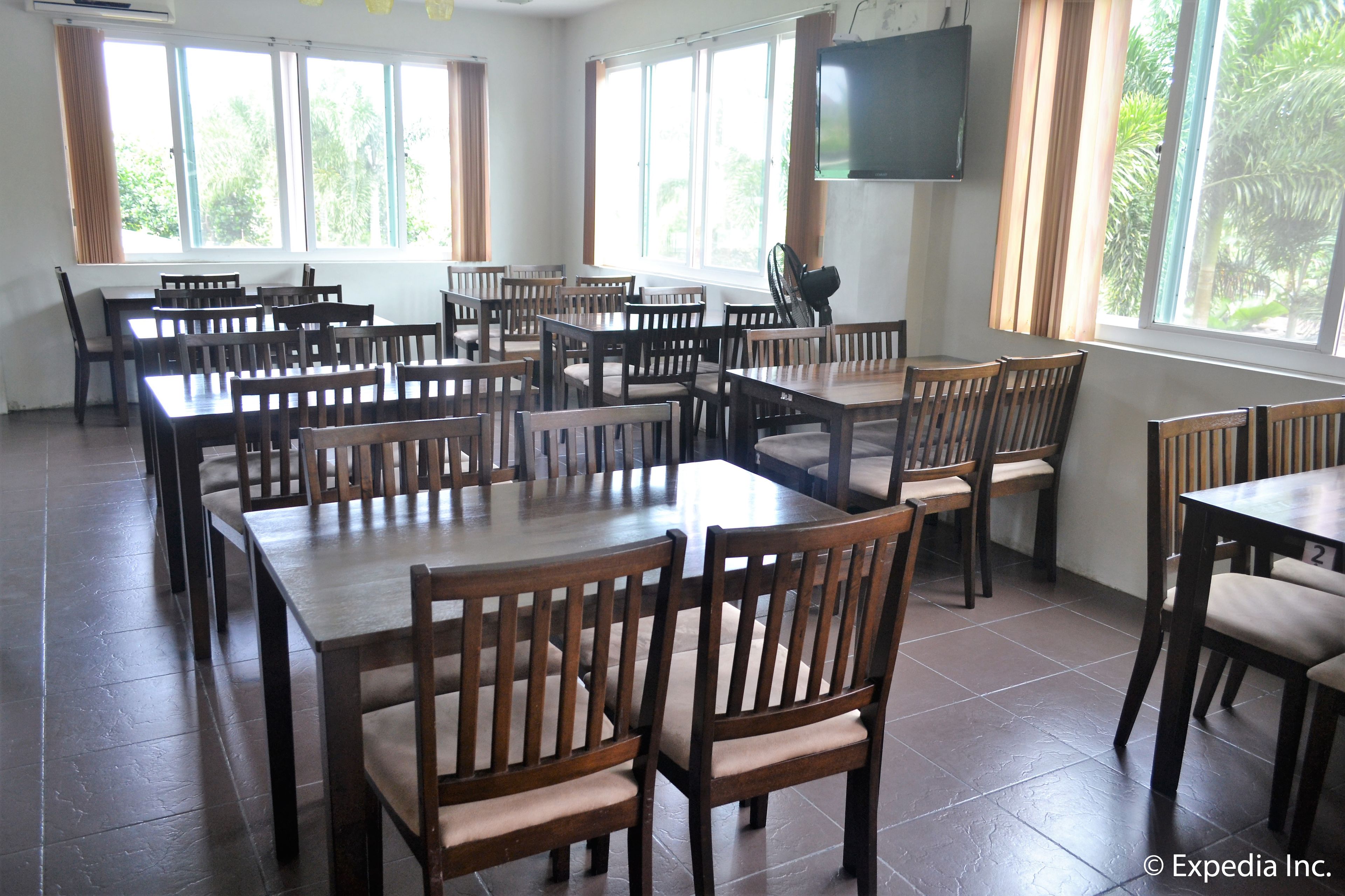 A spacious dining area with multiple wooden tables and chairs, large windows with curtains, and a flat-screen TV mounted on the wall.