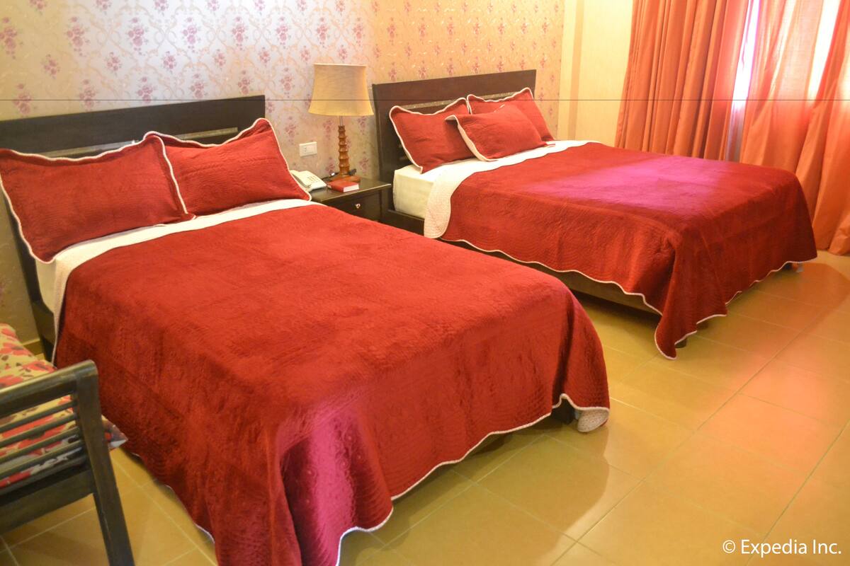 Quarto de hotel com duas camas, cada uma com roupa de cama vermelha e detalhes brancos. Entre as camas, há uma mesa de cabeceira com uma luminária e um telefone. O quarto tem papel de parede floral e piso de azulejos.