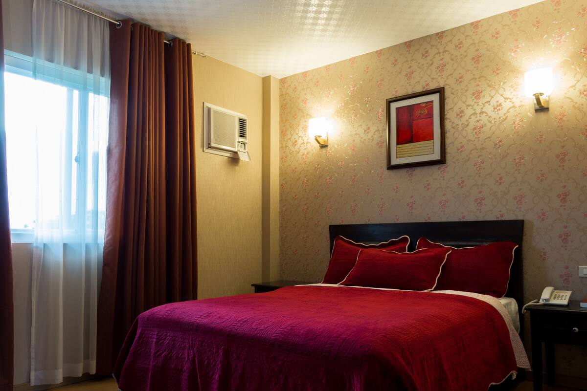 Quarto de hotel com cama, mesa de cabeceira, telefone e luminária na parede.
