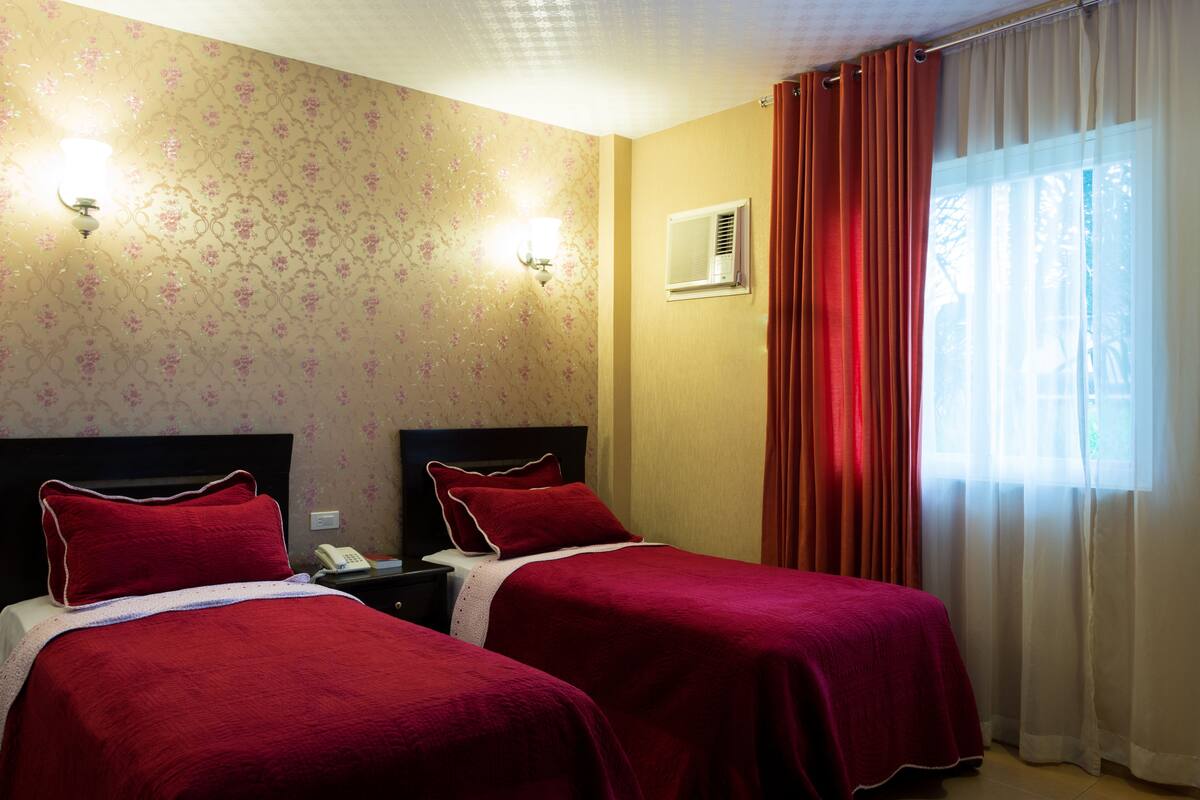 Quarto de hotel com duas camas, papel de parede floral, mesa de cabeceira com telefone e janela com cortinas.