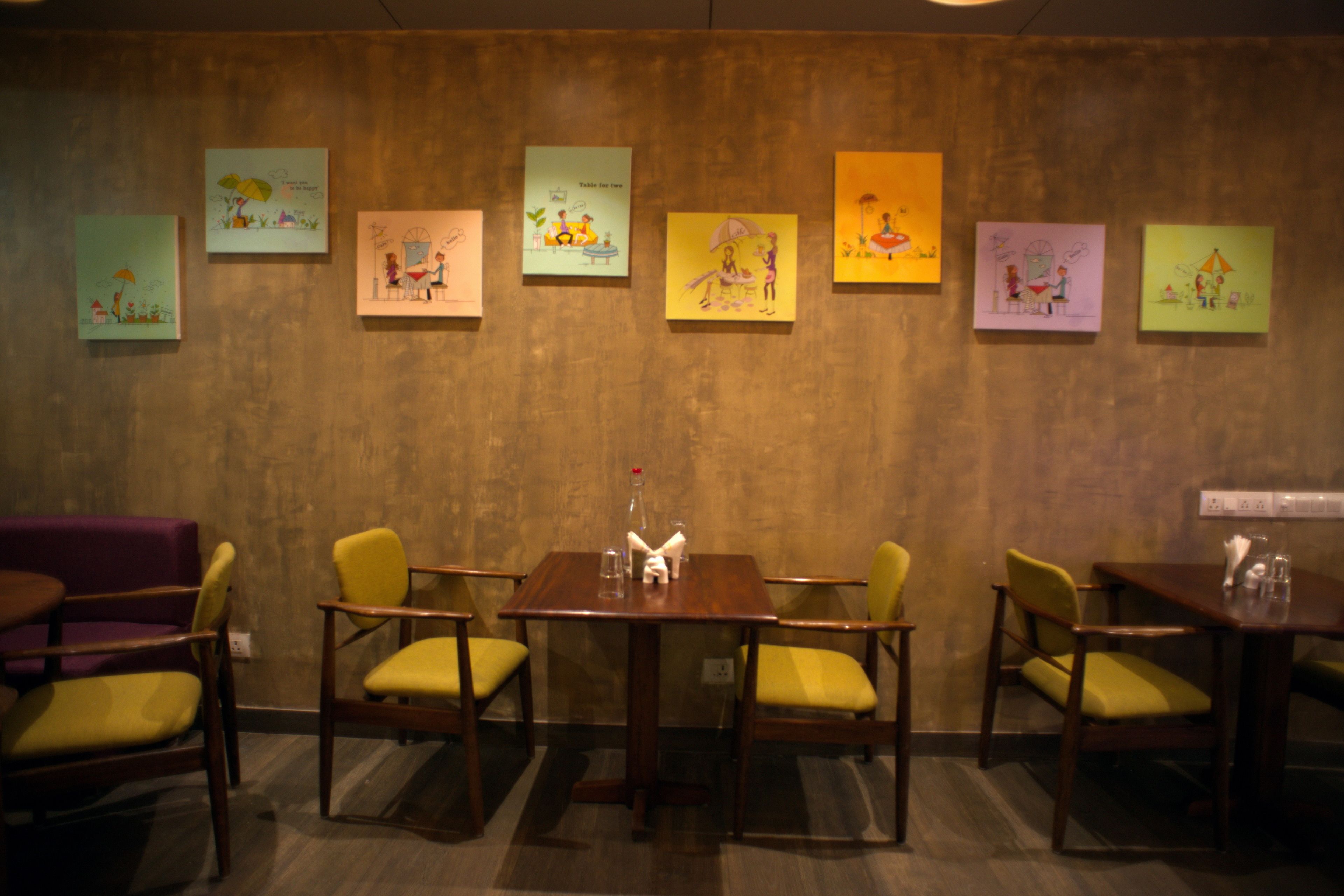 A dining area with a wooden table and yellow chairs, surrounded by six framed artworks on the wall.