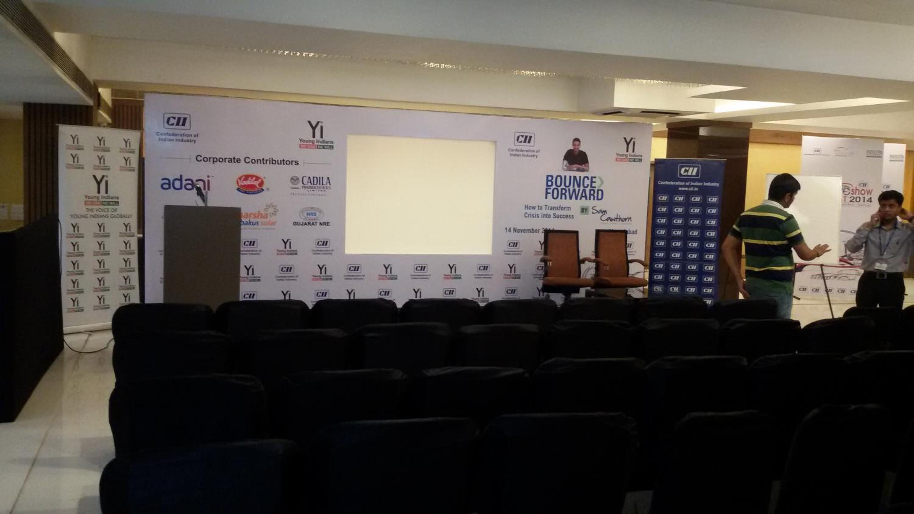 An indoor event space with rows of chairs facing a stage with a backdrop displaying logos and text.