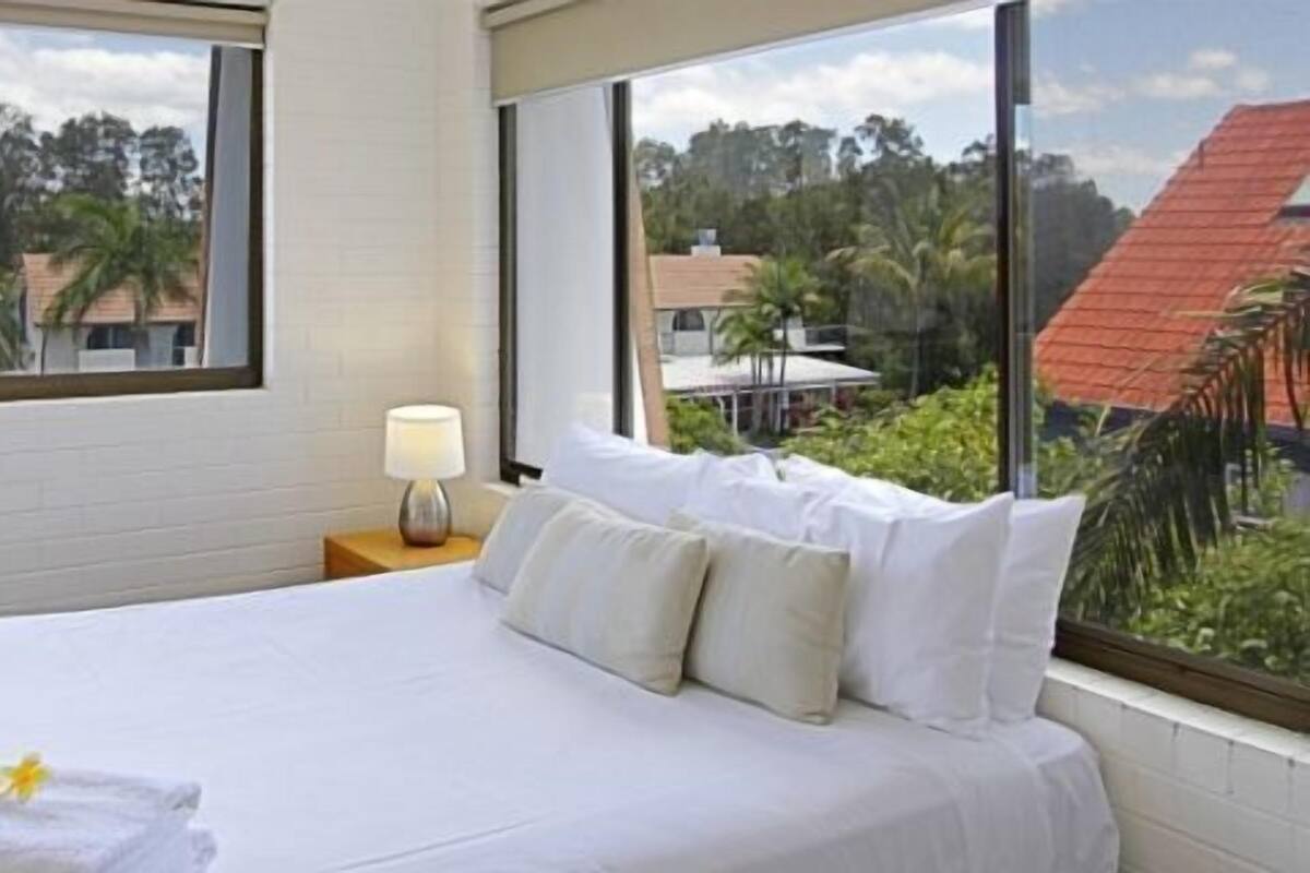 A hotel room with a large bed, a nightstand with a lamp, and a view of a tropical landscape.